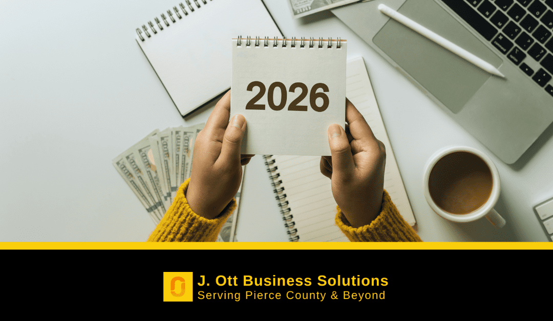 Two hands holding a paper at a desk that reads: 2026. A small stack of money, notebooks, a computer, and cup of coffee are on the desk.